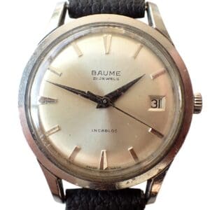 A Baume & Mercier Date 1950s Quality Watch with a round silver case, cream dial, silver markers, date at 3 o’clock, black leather strap, hands at 10:10, and “Baume 21 Jewels Incabloc” on the dial.