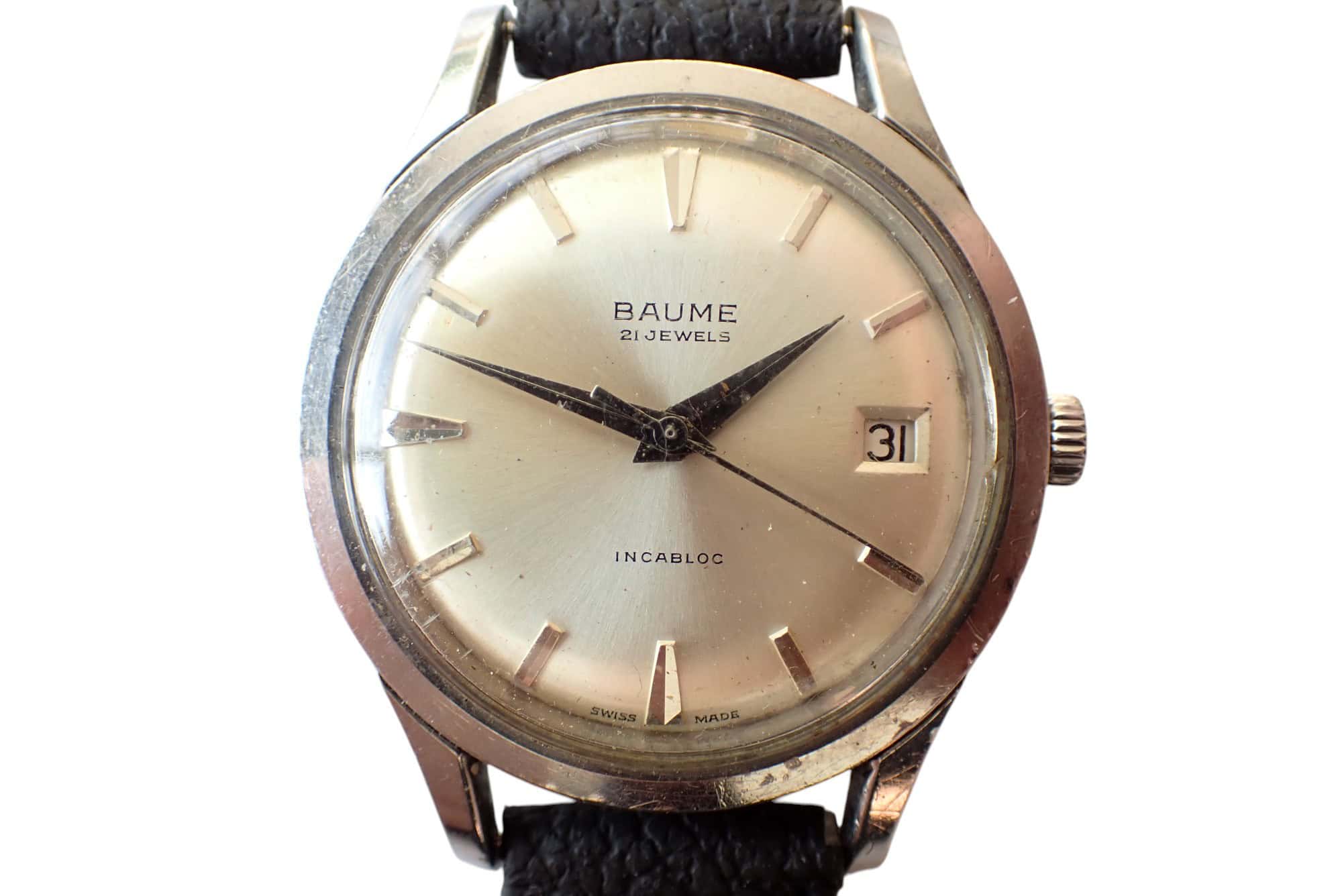 A Baume & Mercier Date 1950s Quality Watch with a round silver case, cream dial, silver markers, date at 3 o’clock, black leather strap, hands at 10:10, and “Baume 21 Jewels Incabloc” on the dial.