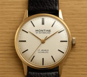 A classic Montine of Switzerland wristwatch with a gold case, white dial, black hour markers, and a black leather strap, placed on a wooden surface. The dial reads 17 JEWELS INCABLOC.