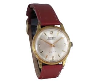 A vintage Nivada Compensamatic wristwatch with a gold-tone case, white dial, and red leather strap. The watch face displays numbers at 3, 6, 9, and 12 o'clock and has a small crown on the right side.