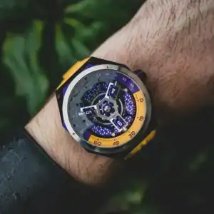 A close-up of a wrist wearing the NUBEO SKYLAB AUTOMATIC LIMITED EDITION Indigo NB-6083-0H, featuring a modern, colourful dial with yellow accents, set against blurred green foliage.