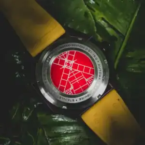 The NUBEO SKYLAB AUTOMATIC LIMITED EDITION Indigo NB-6083-0H wristwatch features a yellow strap and a red case back with a white geometric pattern, shown from behind as it rests on glossy green leaves in low lighting.