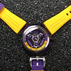 The NUBEO SKYLAB AUTOMATIC LIMITED EDITION Indigo NB-6083-0H features a sporty purple and yellow face with a matching strap. Bold yellow V-shaped strap sections frame the round watch, showcased on black gravel.