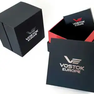 An open black and red Vostok Europe gift box with a booklet lies beside the Vostok Europe Almaz Automatic Line NH35A/320B259. The box and lid display the Vostok Europe logo and branding in metallic lettering.