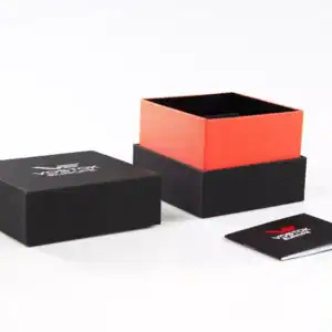 The Vostok Europe Almaz Automatic Line NH35A/320B259 is shown in a black and red square gift box with its black lid off and beside it, along with a small folded black card with white and red text, all set on a white surface.